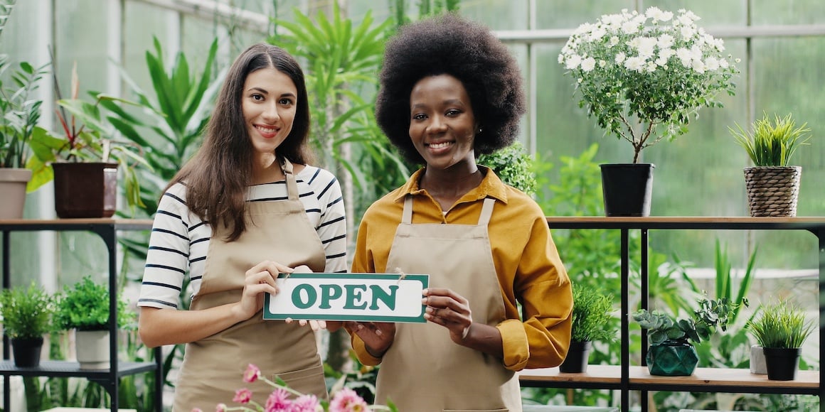 Two smiling female business owners holding an open sign in the florist shop they opened using small business grants
