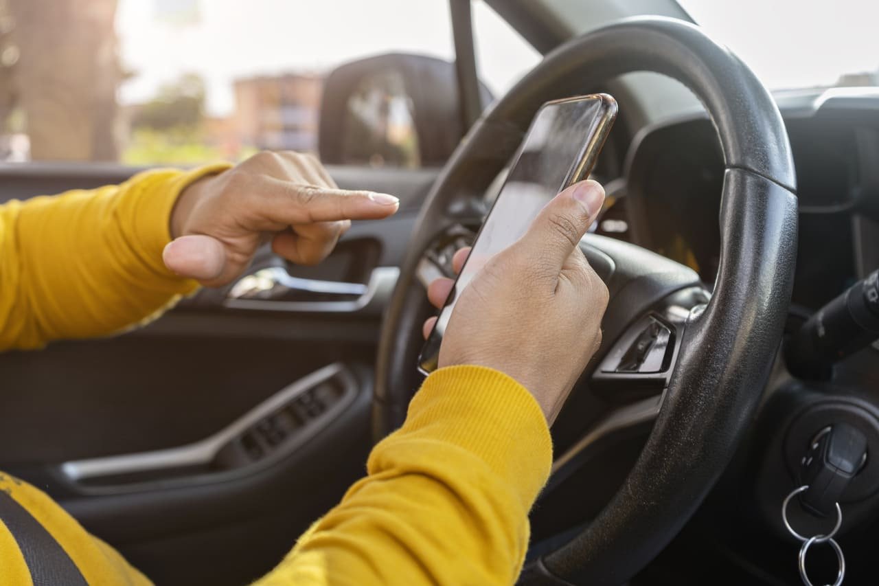 Hands holding a smart phone in drivers seat of car - 4 Best Side Hustles for Additional Income