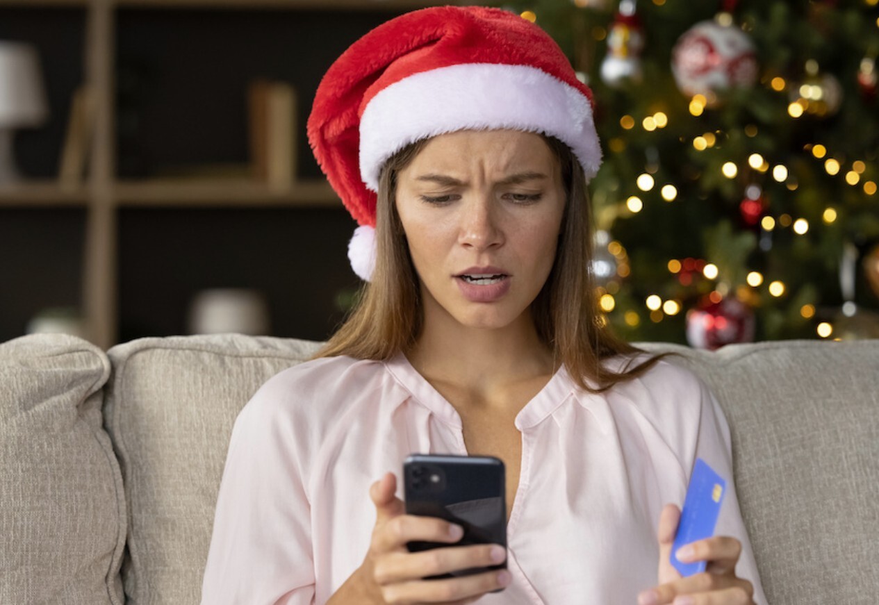 Concerned young woman in a Santa hat holding her debit card and staring at her phone after falling for a holiday scam